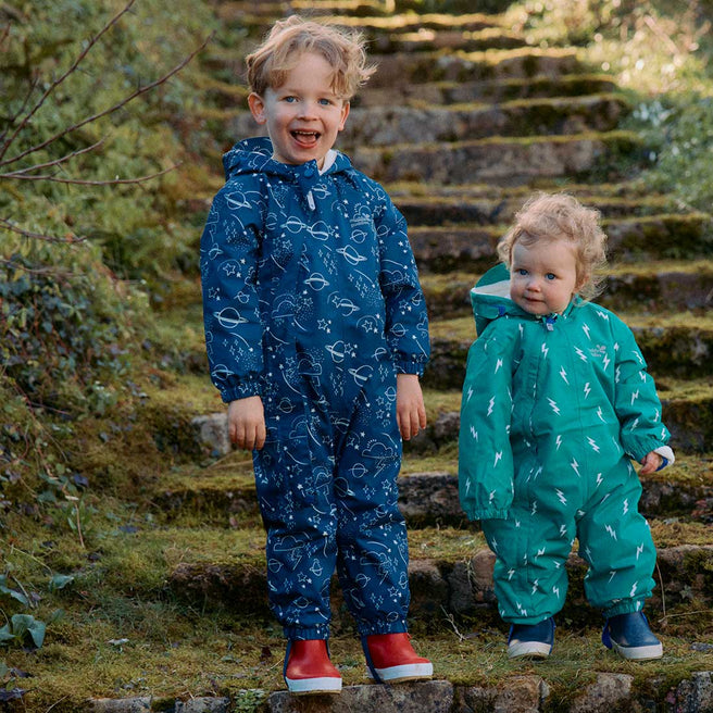 Two children wearing waterproof scampsuits on stone steps. One is a navy '3 in 1 Waterproof Scampsuit Navy' and the other is green.